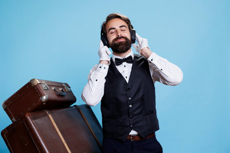 Joyful bellboy dancing and having fun with music against blue background, using audio headset and enjoying songs. Man dressed in suit showing dance moves, feeling confident on camera.の写真素材