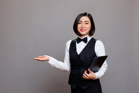 Smiling asian waitress holding digital tablet and welcoming guest with hand portrait. Restaurant receptionist standing with portable device and showing with arm while looking at cameraの写真素材