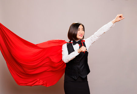 Cheerful asian woman wearing waitress uniform and superwoman cloak while posing in studio. Smiling receptionist raising hand with clenched fist standing in fluttering superhero capeの写真素材