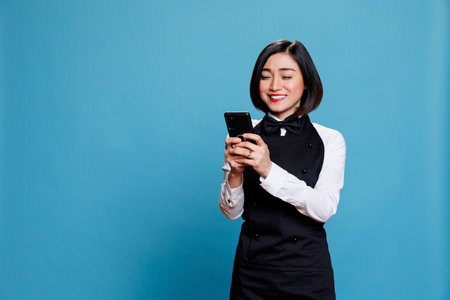 Hotel receptionist holding smartphone and checking message from satisfied customer. Asian waitress with excited face expression reading service positive reviews on mobile phoneの写真素材
