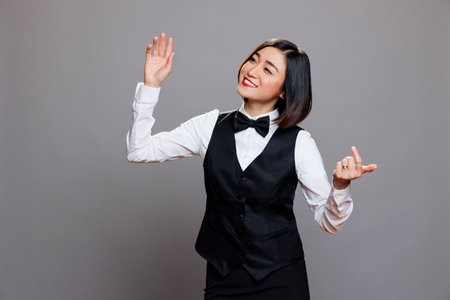 Cheerful asian waitress in uniform laughing and dancing while having fun. Joyful smilng young woman receptionist moving body and hands to music beats while relaxing in studioの写真素材
