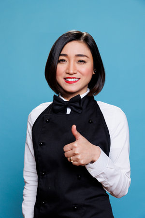 Asian waitress displaying satisfaction with thumb up gesture and looking at camera with cheerful...