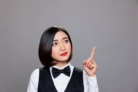 Young attractive asian woman receptionist pointing and looking upwards, showcasing direction. Restaurant waitress wearing uniform showing with finger up while posing in studioの写真素材