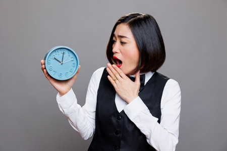 Tired overslept asian waitress in uniform holding circle dial alarm clock and checking time. Restaurant sleepy woman receptionist looking at round retro watch while yawningの写真素材