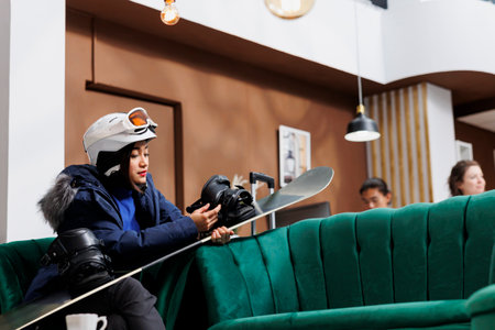 A female traveler securing snowboarding gear and waiting for reservation in hotel reception. Young woman wears winter attire and helmet grasping her snowboard ready for snow activities in ski resort.の写真素材