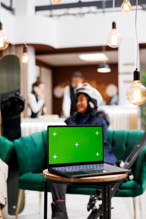 Detailed shot of digital device with isolated copyspace template placed on bench in lounge area of luxury ski mountain resort. Woman in wintersports gear at background of laptop with green screen.の写真素材