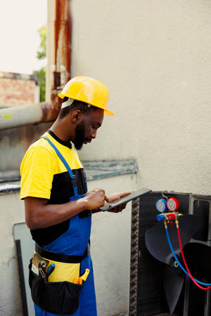 Precise serviceman fixing leaks, replacing filters and other necessary repairs to prevent major breakdowns. African american wireman troubleshooting air conditioner, writing findings on clipboardの写真素材