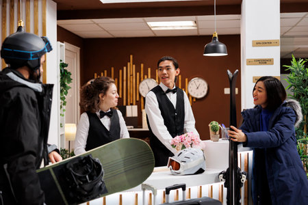 Happy couple in winter gear stands in hotel reception during holiday assisted by receptionists. Visitors with skiing and snowboarding equipment hints at snowy adventures at luxury ski resort.の写真素材