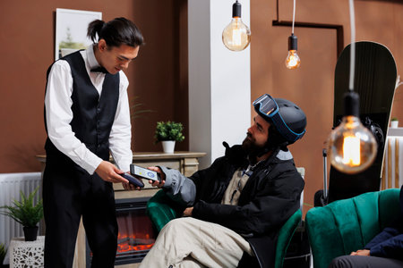 In hotel lobby, male tourist makes contactless payments with smartphone for wintersport gear rental. Caucasian man in snow clothing pays with mobile device on POS terminal held by ski resort employee.の写真素材