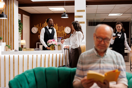 Elderly female tourist with baggage converses with male receptionist at front desk of luxury resort. While elderly guy reads a novel in hotel lobby, senior woman completes booking process at counter.の写真素材