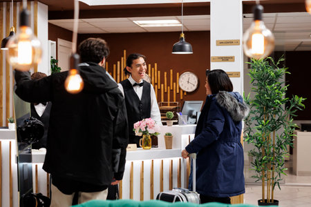 Young couple with winter clothes and suitcase arriving at front desk of luxury holiday resort for room reservation. Friendly concierge welcomes two tourists with ski attire at hotel reception counter.の写真素材
