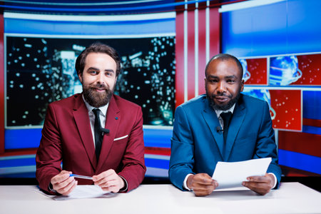 Journalists reporting latest news live on late night show, transmitting information about international events in newsroom. Diverse team of tv hosts working on media entertainment segment.の写真素材