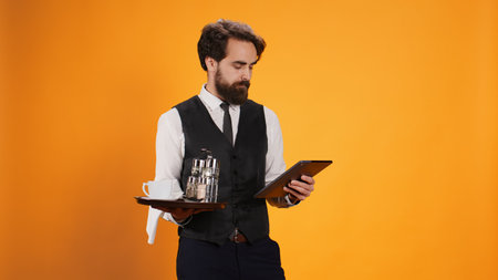 Dedicated staff member calculates check on tablet while working in five star restaurant to serve clients, offer outstanding opulent service. Butler carrying dinner platter and modern gadget.の写真素材