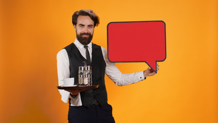 Catering worker presents speech bubble with blank banner space on red icon cutout in studio, indicating advertisement with empty cardboard isolated copyspace. Young butler with food tray.の写真素材
