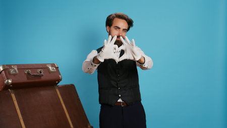 Smiling bellhop does heart shape sign in studio, indicating romantic emotions and flirting on camera while he wears suit and tie. Sweet doorkeeper showing romance gesture for love, sincere feelings.の写真素材
