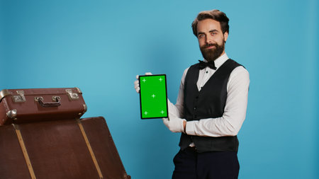 Elegant bellboy holding tablet on camera and presenting blank greenscreen display, symbolizing hotel concierge services with professionalism. Porter in suit and tie pointing at isolated mockup.の写真素材