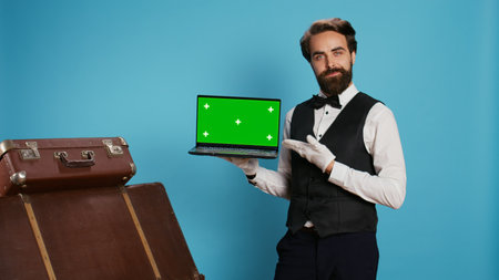 Classy bellhop presents greenscreen on laptop display in studio while he wears gloves next to pile of luggage. Experienced doorman working at luxurious hotel, showing isolated mockup.の写真素材