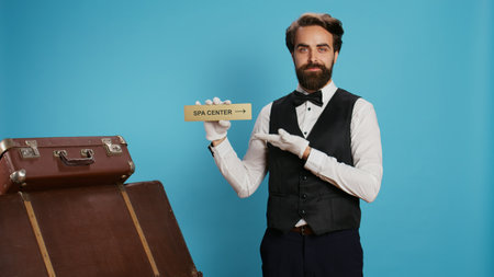 Classy porter shows direction of spa center using indicator, leading tourists to the right way with wall sign. Experienced doorman with white gloves holding pointer to present relaxation area.の写真素材