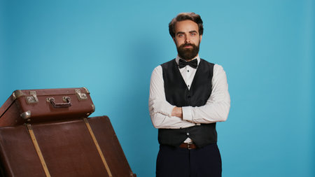 Confident bellboy posing on camera, wearing formal attire while he works at luxurious hotel. Staff member standing next to pile of suitcases and trolley bags, provide excellent concierge service.の写真素材