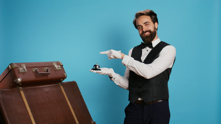 Male employee points at service bell, representing hotel concierge assistance and ringing for help with trolley bags. Formal skilled doorman assisting with travelling luggage, tourism sector.の写真素材