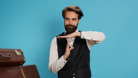 Hotel worker indicates timeout sign in studio, asking for work break and presenting hospitality and travel sector. Stylish doorkeeper in formal clothes showcasing pause symbol with hands.の写真素材