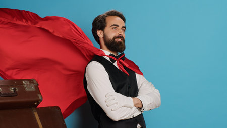 Hotel concierge with red hero cape to help guests with baggage, pretending to be cartoon superhero character on camera. Bellhop acting like fantasy mascot with mantle next to trolley bags.の写真素材
