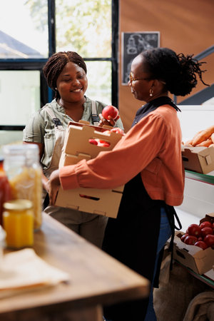 Lady with spectacles receives crates of fresh produce from local farmer at eco friendly store. Storekeeper wearing an apron, carrying boxes of locally grown fruits, purchased from female rancher.の写真素材