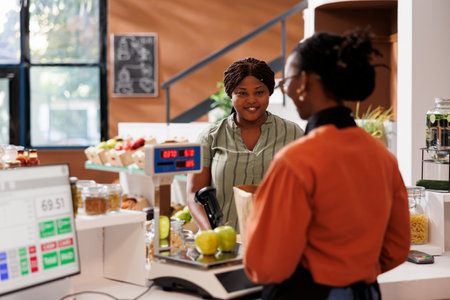 Smiling black lady receiving assistance from African American vendor at the checkout counter with her locally grown produce. Customer at cashier desk in zero waster store purchasing organic products.の写真素材