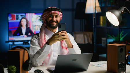 Portrait of smiling Arab freelancer enjoying leisure at home, typing on his smartphone, surfing the net. Happy middle eastern man in traditional attire chatting with friends using mobile phoneの写真素材