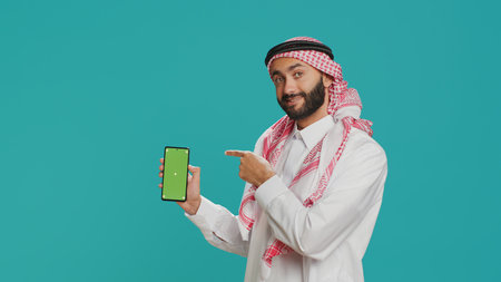 Middle eastern guy shows greenscreen template on smartphone, presenting blank copyspace display on mobile gadget. Young adult in muslim traditional costume points at isolated chromakey.の写真素材