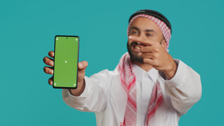 Muslim man on mobile device with greenscreen theme, Putting forward isolated mockup chromakey on smartphone display. Young arabic person exhibits blank copyspace template design.の写真素材