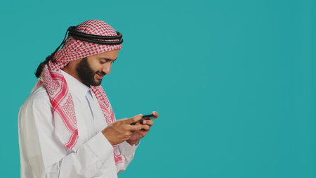 Arabic person playing mobile videogames on smartphone with tensed expression, wears islamic headscarf and thobe. Muslim gamer enjoying online gaming experience on phone app in studio.の写真素材