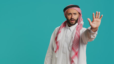 Middle eastern guy doing stop gesture with palm, expressing negativity and refusal in studio. Young male model presenting rejection, being disappointed with stuff over backdrop.の写真素材