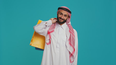 Arab adult carrying shopping bags over blue background, posing with confidence after successful session of buying products. Young person in muslim clothes purchasing goods from mall on sale.の写真素材