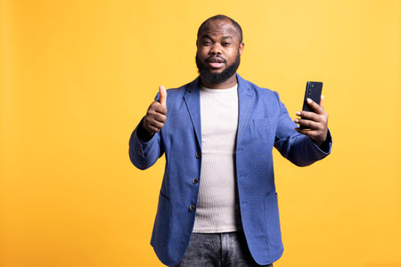 Satisfied african american man doing thumbs up sign, recommending smartphone, isolated over studio background. Pleased BIPOC person doing like hand gesturing while holding mobile phoneの写真素材