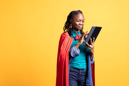 BIPOC girl wearing superhero costume for Halloween, using tablet isolated over studio background. Teenager dressed as comic book hero for event pressing device touchscreenの写真素材
