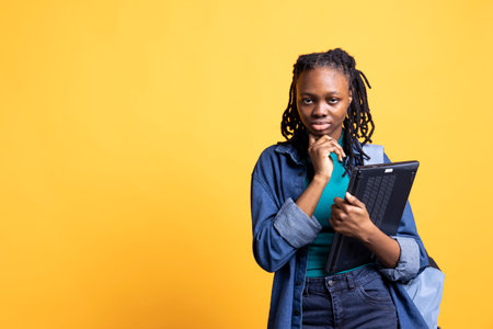 Portrait of pensive woman holding laptop, brainstorming during work shift, isolated over studio background. Employee with notebook doing thinking hand gesturing, ready to do tasksの写真素材