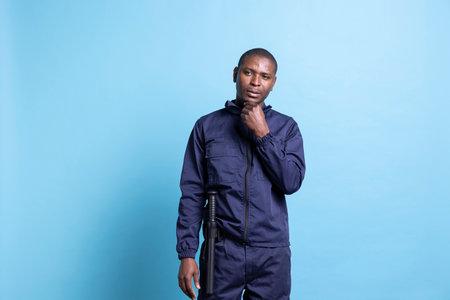 Thoughtful security personnel with uniform and baton thinking of ways to better protect the premises, blue background. Pensive guardian working on safeguarding and securing, patrol service.の写真素材