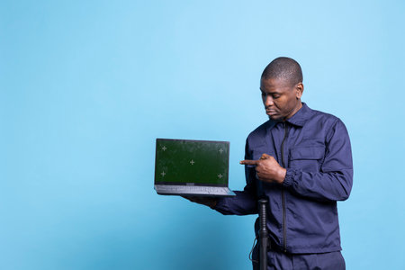 Male security guard pointing at his laptop display with copyspace, showing a greenscreen while he poses on camera. Trained armed officer standing against blue background, patrol service.の写真素材