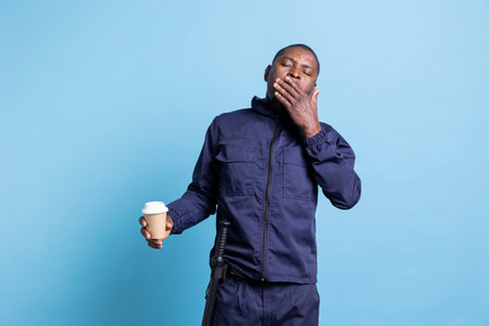 Portrait of african american agent yawning while he enjoys caffeine drink, trying to get over fatigue with a cup of coffee. Male security officer deals with exhaustion from night shifts.の写真素材