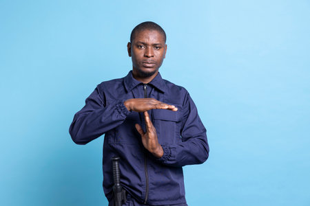 Patrol agent showing a timeout gesture against blue background, asking to take a break while he is on protection duty. Staff in bodyguard uniform showing a pause symbol to relax.の写真素材
