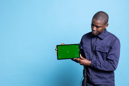 Smiling armed guard holds a greenscreen display on tablet in studio, posing in his uniform against blue background. Young security agent shows an isolated copyspace layout, job duty.の写真素材