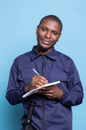 Security officer in work uniform writes important details on notebook, posing against blue background. Smiling proud mall guard taking notes of information to keep evidence of responsibilities.の写真素材