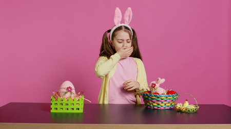 Exhausted little child decorating festive ornaments for easter sunday celebration, creating colorful cute arrangements with painted eggs. Tired girl yawning and falling asleep on the table. Camera B.の写真素材