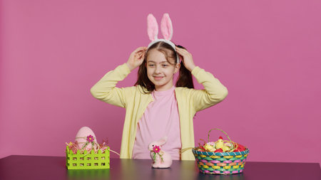 Smiling confident toddler placing bunny ears on her head and waving, saying hello against pink background. Cute joyful child creating decorations for easter sunday holiday event. Camera A.の写真素材
