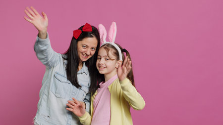 Smiling cheerful mother and little girl waving in front of camera, having fun and enjoying easter holiday celebration. Joyful schoolgirl posing with her mom in studio, saying hello. Camera A.の写真素材