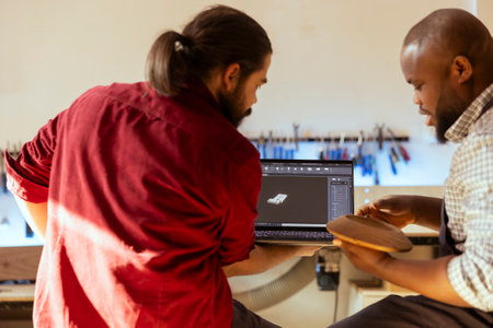 Woodworker looking over blueprints on laptop in CAD software with apprentice. Manufacturer and coworker designing custom 3D furniture on notebook in studio, preparing for customer orderの写真素材