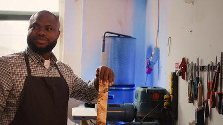 Portrait of jolly man grinning while inspecting wood in furniture assembling shop, satisfied by quality. Joyful woodworking expert in studio evaluates lumber block, pleased by lack of damage, camera Bの写真素材