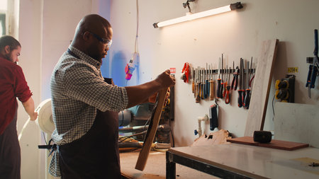 BIPOC carpenter in woodworking shop inspecting lumber piece before assembling furniture, checking for scratches. Cabinetmaker evaluates timber block, picking right tool from rack for it, camera Bの写真素材
