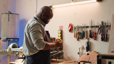 African american artisan in studio using sandpaper for smoothing wooden surface, creating wood art designs. BIPOC person using sanding sheets to refurbish damages suffered by timber block, camera Aの写真素材